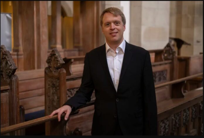 Stefan Donner steht in einem Innenraum einer Kirche. Er hat kurze blonde Haare, trägt einen schwarzen Anzug mit weißem Hemd und lächelt leicht in die Kamera. Mit einer Hand stützt er sich locker auf eine kunstvoll geschnitzte Holzbank. Im Hintergrund sind weitere hölzerne Sitzreihen, Säulen und warme Beleuchtung zu sehen.
