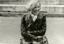 hätt ich nicht Disteln im Herz / Kulturforum Berlin Das Bild ist Schwarz-Weiß-Porträtfoto von Ingeborg Bachmann. Sie sitzt im Freien, trägt einen glänzenden Lackmantel und ein helles Halstuch und lacht. Ihre kinnlangen hellen Haare wehen im Wind. Im Hintergrund sind steinerne Stufen zu sehen.
