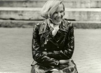 hätt ich nicht Disteln im Herz / Kulturforum Berlin Das Bild ist Schwarz-Weiß-Porträtfoto von Ingeborg Bachmann. Sie sitzt im Freien, trägt einen glänzenden Lackmantel und ein helles Halstuch und lacht. Ihre kinnlangen hellen Haare wehen im Wind. Im Hintergrund sind steinerne Stufen zu sehen.