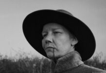 45. Istanbul Film Festival / Kulturforum Istanbul Das Bild zeigt eine Person in einer Schwarz-Weiß-Aufnahme, die im Freien steht. Sie trägt einen breitkrempigen Hut und einen hochgeschlossenen Mantel. Ihr Blick ist ernst und leicht nach vorne gerichtet. Im Hintergrund sind unscharf Gräser oder eine natürliche Landschaft zu erkennen.
