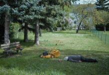European Union Short Film Festival 2026 / Kulturforum Ottawa Das Bild zeigt eine ruhige Gartenszene. Auf einer Wiese liegt eine ältere Person bäuchlings im Gras. In der Nähe steht ein gelber Rasenmäher. Links im Bild befindet sich eine Holzbank mit einer Handtasche darauf. Im Hintergrund sind Bäume, ein Gartenzaun und ein Haus zu sehen. Die Szene wirkt still und leicht rätselhaft.