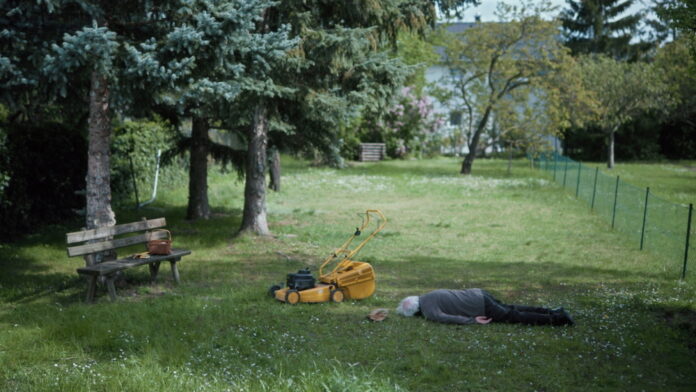 Das Bild zeigt eine ruhige Gartenszene. Auf einer Wiese liegt eine ältere Person bäuchlings im Gras. In der Nähe steht ein gelber Rasenmäher. Links im Bild befindet sich eine Holzbank mit einer Handtasche darauf. Im Hintergrund sind Bäume, ein Gartenzaun und ein Haus zu sehen. Die Szene wirkt still und leicht rätselhaft.