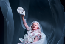 SOUND SCENE: CRYSTN HUNT AKRON / Kulturforum Washington Das Bild zeigt Crystn Hunt Akron in einem auffälligen, skulpturalen Kostüm aus vielen hellen, stäbchenartigen Elementen. In der linken Hand hält sie helle, fließende Stoffbahnen, in der rechten eine bereits zerdrückte Plastikflasche.