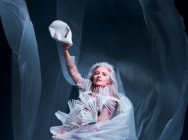 SOUND SCENE: CRYSTN HUNT AKRON / Kulturforum Washington Das Bild zeigt Crystn Hunt Akron in einem auffälligen, skulpturalen Kostüm aus vielen hellen, stäbchenartigen Elementen. In der linken Hand hält sie helle, fließende Stoffbahnen, in der rechten eine bereits zerdrückte Plastikflasche.