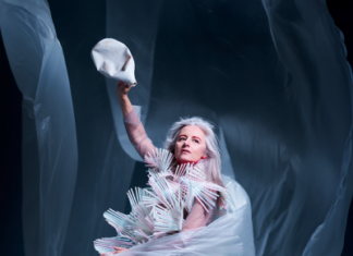 SOUND SCENE: CRYSTN HUNT AKRON / Kulturforum Washington Das Bild zeigt Crystn Hunt Akron in einem auffälligen, skulpturalen Kostüm aus vielen hellen, stäbchenartigen Elementen. In der linken Hand hält sie helle, fließende Stoffbahnen, in der rechten eine bereits zerdrückte Plastikflasche.