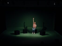 Theater „Lost My Way“ / Kulturforum Washington Das Bild zeigt die Schauspielerin Saskia Norman, die in der Mitte einer Theaterbühne mit dunkelgrünem Hintergrund auf einem schwarzen Klotz sitzt und ihre rechte Hand nach oben hält.