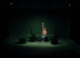 Theater „Lost My Way“ / Kulturforum Washington Das Bild zeigt die Schauspielerin Saskia Norman, die in der Mitte einer Theaterbühne mit dunkelgrünem Hintergrund auf einem schwarzen Klotz sitzt und ihre rechte Hand nach oben hält.