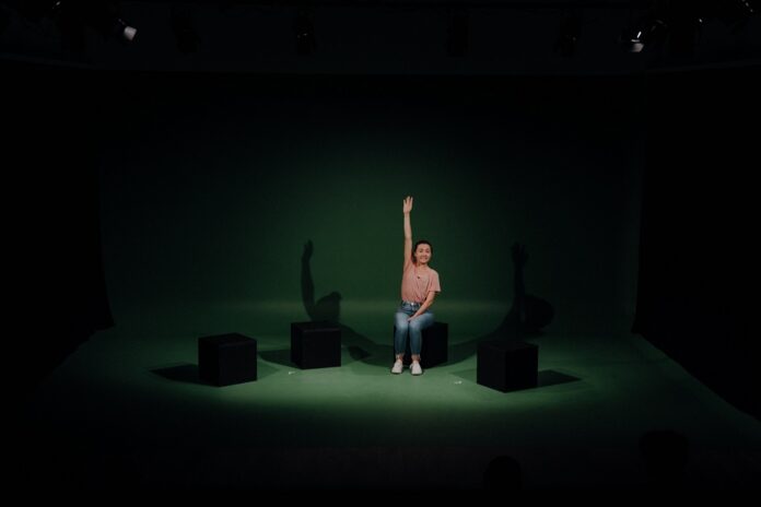 Das Bild zeigt die Schauspielerin Saskia Norman, die in der Mitte einer Theaterbühne mit dunkelgrünem Hintergrund auf einem schwarzen Klotz sitzt und ihre rechte Hand nach oben hält.