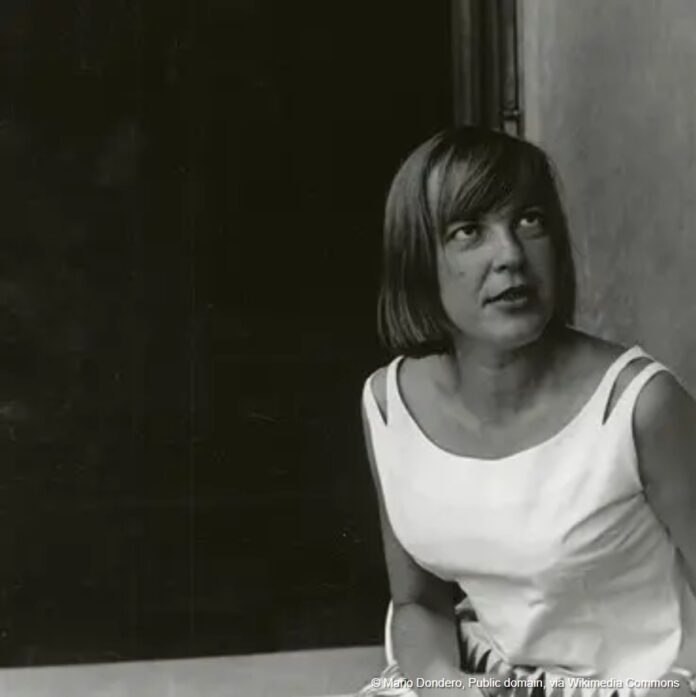 Schwarzweißporträt der Schriftstellerin Ingeborg Bachmann. Sie sitzt seitlich vor einem dunklen Hintergrund und trägt ein ärmelloses, helles Kleid. Ihre Frisur, ein kurzer, glatter Bob umrahmt das Gesicht. Bachmann blickt nach oben rechts außerhalb des Bildausschnitts, ihr Ausdruck wirkt nachdenklich und konzentriert.