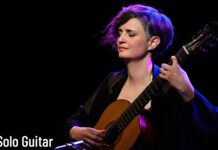 Zsófia Boros / Kulturforum Istanbul Das Bild zeigt die Gitarristin auf einer Bühne, mit geschlossenen Augen ein klassisches Gitarrenstück spielend. Warmes Bühnenlicht in violetten und blauen Tönen hebt sie vor dunklem Hintergrund hervor.