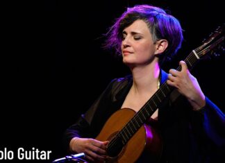 Zsófia Boros / Kulturforum Istanbul Das Bild zeigt die Gitarristin auf einer Bühne, mit geschlossenen Augen ein klassisches Gitarrenstück spielend. Warmes Bühnenlicht in violetten und blauen Tönen hebt sie vor dunklem Hintergrund hervor.