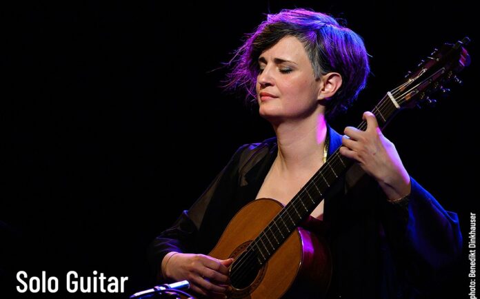 Das Bild zeigt die Gitarristin auf einer Bühne, mit geschlossenen Augen ein klassisches Gitarrenstück spielend. Warmes Bühnenlicht in violetten und blauen Tönen hebt sie vor dunklem Hintergrund hervor.