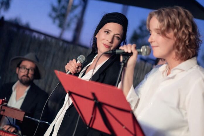 Das Bild zeigt zwei Performerinnen mit Mikrofon und einen Klavierspieler im Hintergrund