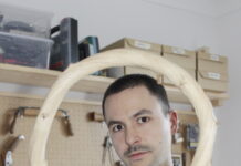 Next! Young European Design / Kulturforum Berlin Das Bild zeigt Jakob Niemann in einer Werkstatt, wie er einen gebogenen Holzbogen in die Kamera hält, der seinen Kopf wie einen Rahmen umgibt. Er hat sehr kurze dunkle Haare und trägt eine graue Arbeitsjacke. Er schaut direkt und ruhig in die Linse. Im Hintergrund sind Werkzeuge ordentlich an einer Lochplatte aufgehängt.