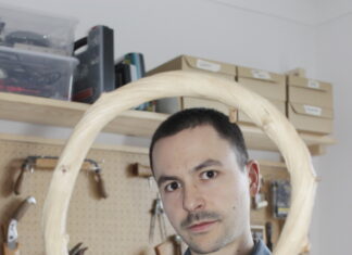 Next! Young European Design / Kulturforum Berlin Das Bild zeigt Jakob Niemann in einer Werkstatt, wie er einen gebogenen Holzbogen in die Kamera hält, der seinen Kopf wie einen Rahmen umgibt. Er hat sehr kurze dunkle Haare und trägt eine graue Arbeitsjacke. Er schaut direkt und ruhig in die Linse. Im Hintergrund sind Werkzeuge ordentlich an einer Lochplatte aufgehängt.