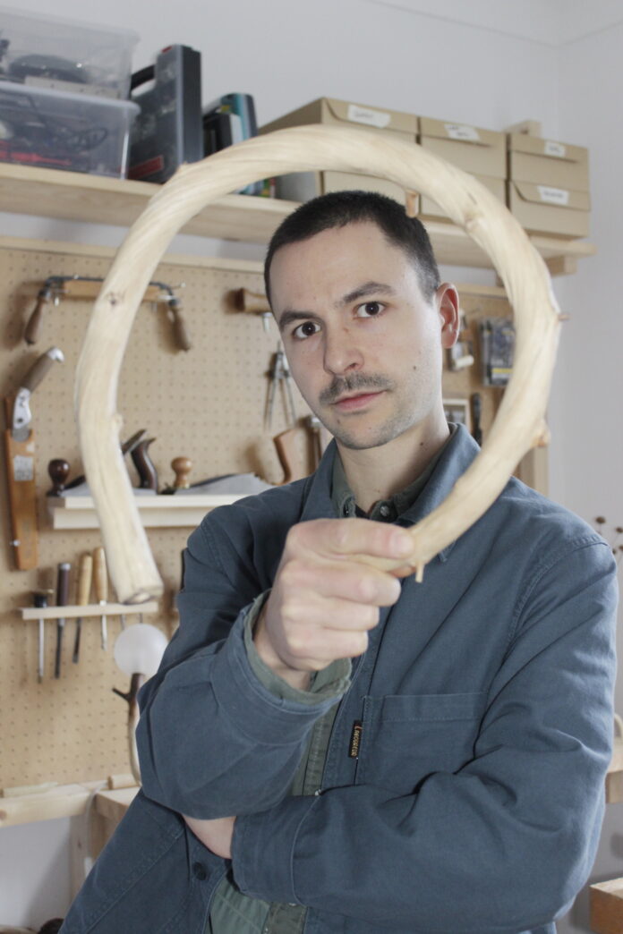 Das Bild zeigt Jakob Niemann in einer Werkstatt, wie er einen gebogenen Holzbogen in die Kamera hält, der seinen Kopf wie einen Rahmen umgibt. Er hat sehr kurze dunkle Haare und trägt eine graue Arbeitsjacke. Er schaut direkt und ruhig in die Linse. Im Hintergrund sind Werkzeuge ordentlich an einer Lochplatte aufgehängt.