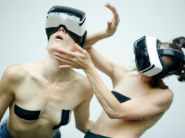 AnotherR – Performance / Kulturforum Bern Das Bild zeigt zwei weibliche Personen tragen VR-Brillen und bewegen sich performativ vor einem weißen Hintergrund. Ihr Oberkörper ist dabei frei und auf ihren Brüsten kleben schwarze Tapes.