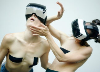 AnotherR – Performance / Kulturforum Bern Das Bild zeigt zwei weibliche Personen tragen VR-Brillen und bewegen sich performativ vor einem weißen Hintergrund. Ihr Oberkörper ist dabei frei und auf ihren Brüsten kleben schwarze Tapes.