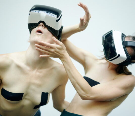 AnotherR – Performance / Kulturforum Bern Das Bild zeigt zwei weibliche Personen tragen VR-Brillen und bewegen sich performativ vor einem weißen Hintergrund. Ihr Oberkörper ist dabei frei und auf ihren Brüsten kleben schwarze Tapes.