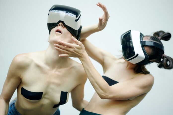 Das Bild zeigt zwei weibliche Personen tragen VR-Brillen und bewegen sich performativ vor einem weißen Hintergrund. Ihr Oberkörper ist dabei frei und auf ihren Brüsten kleben schwarze Tapes.