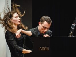 Gugg Piano Duo / Kulturforum New York Das Bild zeigt das Klavierduo The Gugg Piano Duo, bestehend aus Tereza Gugg-Kalabova und Johannes Gugg, während einer intensiven Konzertperformance. Links ist Tereza Gugg-Kalabova zu sehen: Ihr lockiges Haar fliegt in Bewegung nach oben, was die Energie ihres Spiels sichtbar macht. Mit geschlossenen Augen und einem ausdrucksstarken Gesicht wirkt sie ganz in die Musik vertieft. Sie trägt ein elegantes schwarzes Konzertkleid. Rechts sitzt Johannes Gugg am Flügel. Er ist leicht nach vorne geneigt, mit konzentriertem Blick und gespannter Körperhaltung, ganz auf die Ausführung fokussiert. Sein Ausdruck vermittelt Präzision und Hingabe. Beide spielen gemeinsam an einem Konzertflügel und wirken dabei stark aufeinander abgestimmt. Die Szene zeigt eindrucksvoll das Zusammenspiel und die künstlerische Intensität des Duos in einem Moment voller Ausdruck und Dynamik.
