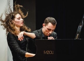 Gugg Piano Duo / Kulturforum New York Das Bild zeigt das Klavierduo The Gugg Piano Duo, bestehend aus Tereza Gugg-Kalabova und Johannes Gugg, während einer intensiven Konzertperformance. Links ist Tereza Gugg-Kalabova zu sehen: Ihr lockiges Haar fliegt in Bewegung nach oben, was die Energie ihres Spiels sichtbar macht. Mit geschlossenen Augen und einem ausdrucksstarken Gesicht wirkt sie ganz in die Musik vertieft. Sie trägt ein elegantes schwarzes Konzertkleid. Rechts sitzt Johannes Gugg am Flügel. Er ist leicht nach vorne geneigt, mit konzentriertem Blick und gespannter Körperhaltung, ganz auf die Ausführung fokussiert. Sein Ausdruck vermittelt Präzision und Hingabe. Beide spielen gemeinsam an einem Konzertflügel und wirken dabei stark aufeinander abgestimmt. Die Szene zeigt eindrucksvoll das Zusammenspiel und die künstlerische Intensität des Duos in einem Moment voller Ausdruck und Dynamik.