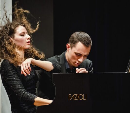 Gugg Piano Duo / Kulturforum New York Das Bild zeigt das Klavierduo The Gugg Piano Duo, bestehend aus Tereza Gugg-Kalabova und Johannes Gugg, während einer intensiven Konzertperformance. Links ist Tereza Gugg-Kalabova zu sehen: Ihr lockiges Haar fliegt in Bewegung nach oben, was die Energie ihres Spiels sichtbar macht. Mit geschlossenen Augen und einem ausdrucksstarken Gesicht wirkt sie ganz in die Musik vertieft. Sie trägt ein elegantes schwarzes Konzertkleid. Rechts sitzt Johannes Gugg am Flügel. Er ist leicht nach vorne geneigt, mit konzentriertem Blick und gespannter Körperhaltung, ganz auf die Ausführung fokussiert. Sein Ausdruck vermittelt Präzision und Hingabe. Beide spielen gemeinsam an einem Konzertflügel und wirken dabei stark aufeinander abgestimmt. Die Szene zeigt eindrucksvoll das Zusammenspiel und die künstlerische Intensität des Duos in einem Moment voller Ausdruck und Dynamik.