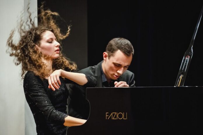 Das Bild zeigt das Klavierduo The Gugg Piano Duo, bestehend aus Tereza Gugg-Kalabova und Johannes Gugg, während einer intensiven Konzertperformance. Links ist Tereza Gugg-Kalabova zu sehen: Ihr lockiges Haar fliegt in Bewegung nach oben, was die Energie ihres Spiels sichtbar macht. Mit geschlossenen Augen und einem ausdrucksstarken Gesicht wirkt sie ganz in die Musik vertieft. Sie trägt ein elegantes schwarzes Konzertkleid. Rechts sitzt Johannes Gugg am Flügel. Er ist leicht nach vorne geneigt, mit konzentriertem Blick und gespannter Körperhaltung, ganz auf die Ausführung fokussiert. Sein Ausdruck vermittelt Präzision und Hingabe. Beide spielen gemeinsam an einem Konzertflügel und wirken dabei stark aufeinander abgestimmt. Die Szene zeigt eindrucksvoll das Zusammenspiel und die künstlerische Intensität des Duos in einem Moment voller Ausdruck und Dynamik.