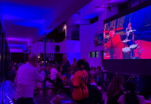 Veranstaltung: ESC Watch Party / Kulturforum Accra Ein großer Innenraum mit gedämpftem violettem und blauem Licht. Mehrere Menschen sitzen an Tischen oder stehen zusammen und schauen auf eine sehr große Leinwand an der rechten Seite des Raumes. Auf der Leinwand läuft eine Musik- oder Bühnenshow mit Musiker und Musikerinnen in bunten Kostümen. Im Vordergrund steht eine Person in einem weißen Hemd mit dem Rücken zur Kamera. Daneben sitzt eine weitere Person mit gemusterter Kleidung. In der Mitte des Bildes steht eine Person in einem orangefarbenen Fußballtrikot mit der Nummer 20 und hält Getränke in der Hand. Der Raum wirkt modern und offen, mit hohen Decken, Ventilatoren und langen Fensterfronten auf der linken Seite. Die Atmosphäre erinnert an eine öffentliche Veranstaltung oder ein gemeinsames Public Viewing in einer Bar.