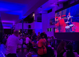 Veranstaltung: ESC Watch Party / Kulturforum Accra Ein großer Innenraum mit gedämpftem violettem und blauem Licht. Mehrere Menschen sitzen an Tischen oder stehen zusammen und schauen auf eine sehr große Leinwand an der rechten Seite des Raumes. Auf der Leinwand läuft eine Musik- oder Bühnenshow mit Musiker und Musikerinnen in bunten Kostümen. Im Vordergrund steht eine Person in einem weißen Hemd mit dem Rücken zur Kamera. Daneben sitzt eine weitere Person mit gemusterter Kleidung. In der Mitte des Bildes steht eine Person in einem orangefarbenen Fußballtrikot mit der Nummer 20 und hält Getränke in der Hand. Der Raum wirkt modern und offen, mit hohen Decken, Ventilatoren und langen Fensterfronten auf der linken Seite. Die Atmosphäre erinnert an eine öffentliche Veranstaltung oder ein gemeinsames Public Viewing in einer Bar.