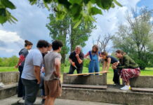 Theater, Film, Holzarbeit / Kulturforum Belgrad Das Bild zeigt die Studierenden beim Holz-Workshop im Freien. Sie stehen im Kreis um Ludwig Wüst, der eine Säge in der Hand hält und etwas erklärt.