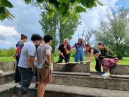 Theater, Film, Holzarbeit / Kulturforum Belgrad Das Bild zeigt die Studierenden beim Holz-Workshop im Freien. Sie stehen im Kreis um Ludwig Wüst, der eine Säge in der Hand hält und etwas erklärt.