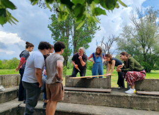 Theater, Film, Holzarbeit / Kulturforum Belgrad Das Bild zeigt die Studierenden beim Holz-Workshop im Freien. Sie stehen im Kreis um Ludwig Wüst, der eine Säge in der Hand hält und etwas erklärt.