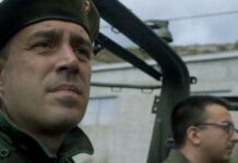 Krava (The Cow) – Pavo Marinković / Kulturforum Ottawa Das Bild zeigt einen Mann in Militäruniform, der in einem offenen Fahrzeug sitzt und ernst nach vorne blickt. Im Hintergrund ist eine zweite uniformierte Person mit Brille zu sehen. Die Szene wirkt angespannt.