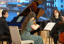 Amatis Trio / Kulturforum Peking Das Bild zeigt die drei Musikerinnen und Musiker bei einem Konzert. Eine Violinistin, ein Cellist und ein Pianist spielen gemeinsam auf einer Bühne vor einem großen Flügel. Im Hintergrund ist eine projizierte Landschaft von Österreich zu sehen.