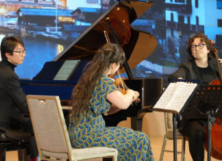 Amatis Trio / Kulturforum Peking Das Bild zeigt die drei Musikerinnen und Musiker bei einem Konzert. Eine Violinistin, ein Cellist und ein Pianist spielen gemeinsam auf einer Bühne vor einem großen Flügel. Im Hintergrund ist eine projizierte Landschaft von Österreich zu sehen.