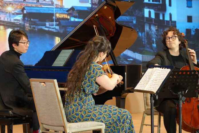 Das Bild zeigt die drei Musikerinnen und Musiker bei einem Konzert. Eine Violinistin, ein Cellist und ein Pianist spielen gemeinsam auf einer Bühne vor einem großen Flügel. Im Hintergrund ist eine projizierte Landschaft von Österreich zu sehen.