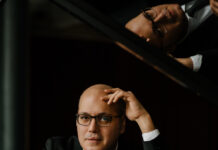 Konzert von Gottlieb Wallisch/ Kulturforum Sarajewo Das Bild zeigt einen Mann mit Brille, in schwarzem Anzug und weißem Hemd, am Klavier sitzend.