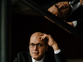 Konzert von Gottlieb Wallisch/ Kulturforum Sarajewo Das Bild zeigt einen Mann mit Brille, in schwarzem Anzug und weißem Hemd, am Klavier sitzend.