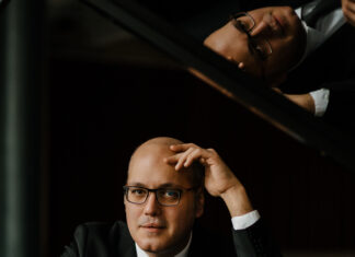 Konzert von Gottlieb Wallisch/ Kulturforum Sarajewo Das Bild zeigt einen Mann mit Brille, in schwarzem Anzug und weißem Hemd, am Klavier sitzend.