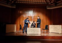 Stefan Zweig Woche / Botschaft Lima Das Bild zeigt eine szenische Lesung auf einer Bühne mit holzverkleideter Rückwand. Drei Schauspieler in dunklen Anzügen stehen beziehungsweise sitzen um einen niedrigen Tisch mit einem Schachbrett. Zwei schwarze Stühle flankieren die Szene. Einer der Darsteller sitzt konzentriert am Tisch, während die beiden anderen mit Manuskriptseiten in der Hand agieren. Die Beleuchtung richtet den Fokus auf die Figuren und erzeugt eine intensive, dramatische Atmosphäre. Die minimalistische Bühnenausstattung unterstreicht den psychologischen Charakter der Aufführung von Stefan Zweigs „Schachnovelle“.
