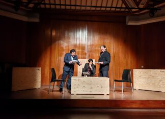 Stefan Zweig Woche / Botschaft Lima Das Bild zeigt eine szenische Lesung auf einer Bühne mit holzverkleideter Rückwand. Drei Schauspieler in dunklen Anzügen stehen beziehungsweise sitzen um einen niedrigen Tisch mit einem Schachbrett. Zwei schwarze Stühle flankieren die Szene. Einer der Darsteller sitzt konzentriert am Tisch, während die beiden anderen mit Manuskriptseiten in der Hand agieren. Die Beleuchtung richtet den Fokus auf die Figuren und erzeugt eine intensive, dramatische Atmosphäre. Die minimalistische Bühnenausstattung unterstreicht den psychologischen Charakter der Aufführung von Stefan Zweigs „Schachnovelle“.