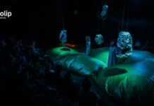 Österreichisches Theater / Botschaft Pristina Das Bild zeigt ein futuristisches Bühnenbild in grün-blau-schwarz mit zwei maskierten Schauspielerinnen.