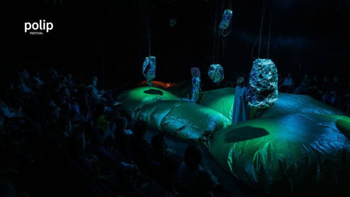 Das Bild zeigt ein futuristisches Bühnenbild in grün-blau-schwarz mit zwei maskierten Schauspielerinnen.
