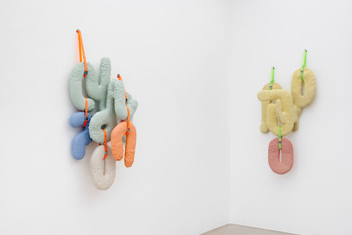 Das Bild zeigt zwei abstrakte keramische Wandskulpturen von Beate Gatschelhofer, bestehend aus pastellfarbenen, strukturierten Formen, die mit farbigen Schnüren an weißen Galeriewänden befestigt sind.