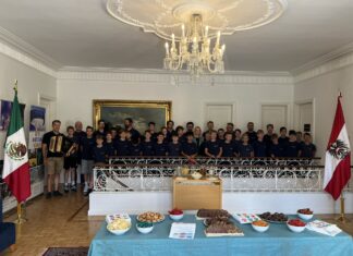 St. Florianer Sängerknaben / Kulturforum Mexiko Die St. Florianer Sängerknaben stehen in einem festlichen Raum mit Kronleuchter, Möbeln und Teppichen. Im Hintergrund sind die Flaggen Österreichs und Mexikos zu sehen.