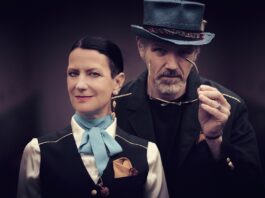 Orlando Trip / Kulturforum New York Eine Frau im Vordergrund trägt eine schwarze Weste über einer weißen Bluse mit hellblauer Schleife und einem goldenen Einstecktuch; ihr Blick ist ruhig und direkt in die Kamera gerichtet. Hinter ihr steht ein Mann mit grauem Zylinder und dunklem Jackett, der eine Brille in der Hand hält und leicht zur Seite schaut. Der unscharfe violett-graue Hintergrund verleiht dem Bild eine theatralische, elegante Atmosphäre.