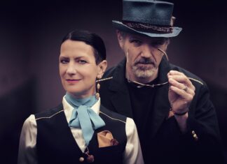 Orlando Trip / Kulturforum New York Eine Frau im Vordergrund trägt eine schwarze Weste über einer weißen Bluse mit hellblauer Schleife und einem goldenen Einstecktuch; ihr Blick ist ruhig und direkt in die Kamera gerichtet. Hinter ihr steht ein Mann mit grauem Zylinder und dunklem Jackett, der eine Brille in der Hand hält und leicht zur Seite schaut. Der unscharfe violett-graue Hintergrund verleiht dem Bild eine theatralische, elegante Atmosphäre.