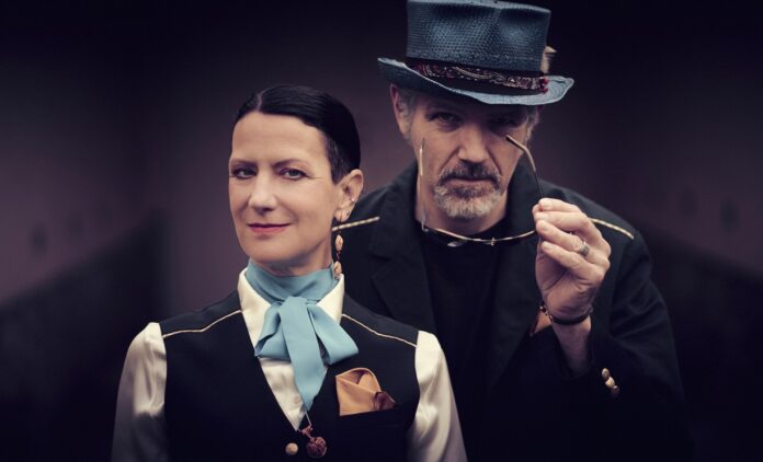 Eine Frau im Vordergrund trägt eine schwarze Weste über einer weißen Bluse mit hellblauer Schleife und einem goldenen Einstecktuch; ihr Blick ist ruhig und direkt in die Kamera gerichtet. Hinter ihr steht ein Mann mit grauem Zylinder und dunklem Jackett, der eine Brille in der Hand hält und leicht zur Seite schaut. Der unscharfe violett-graue Hintergrund verleiht dem Bild eine theatralische, elegante Atmosphäre.