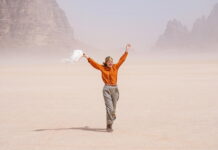 Sotto le Stelle dell’Austria / Kulturforum Rom Das Bild zeigt Schauspielerin Vicky Krieps in ihrer Rolle als Ingeborg Bachmann im Film „Reise in die Wüste“. Sie geht über hellen Wüstensand. Sie trägt eine orangefarbene Bluse, eine beige Hose und ein buntes Kopftuch. In der linken Hand hält sie einen weißen Schal, der im Wind weht. Lachend wirft sie die Arme nach oben. Im Hintergrund sind Felsen zu sehen.